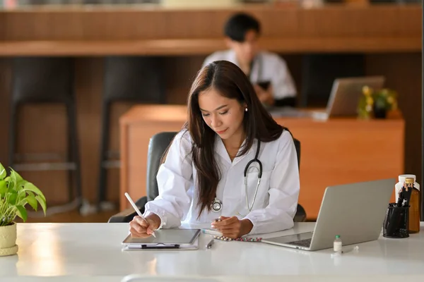 一位年轻的女医生在数码平板电脑上工作 在医院的办公室里 一位身穿白衣的医生正在阅读一份过去的病史研究报告 — 图库照片