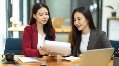 İki çalışkan genç ve zeki iş kadını finansal belgeler üzerinde birlikte çalışıyor ve modern ofiste sonuçları okuyorlar..