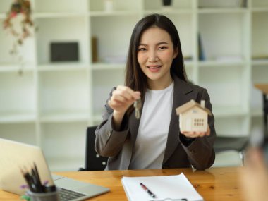 Ev anahtarı ve ev modeliyle Asyalı emlakçı kadın modern bir ofiste oturuyor..