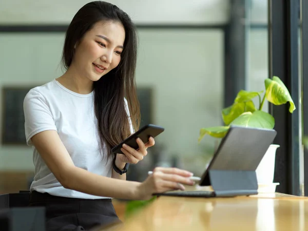 美丽的亚洲女自由职业者在咖啡店用手机和便携式平板电脑工作 — 图库照片
