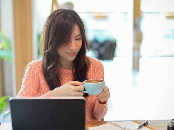 仕事中にカフェでコーヒーを飲むピンクのセーターの美しい若い女性 彼女のポータブルタブレットコンピュータ上でオンライン研究 — ストック写真