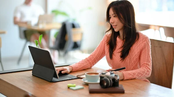 居心地の良いカフェでポータブルタブレットコンピュータで作業ピンクのセーターで魅力的なアジアの若い女性のフリーランサー テーブルの上にコーヒーカップとヴィンテージカメラ — ストック写真