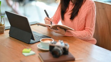 Kafedeki tablet bilgisayarda çalışırken, kadın yazarın çalışma sürecini not defterine planlarken kesilen görüntüsü.