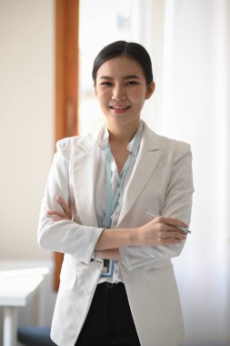 Başarılı ve kendinden emin beyaz takım elbiseli Asyalı genç bir iş kadınının portresi modern ofiste kollarını kavuşturmuş duruyor..