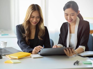 Happy and cheerful asian young business women looking at tablet screen and thinking a creative ideas together.