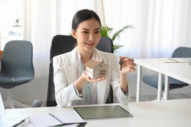 Gorgeous asian young real estate agent holding a house key and house model in minimal office room.