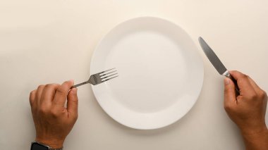 Top view image of a man holding fork and knife over dining table. Blank white ceramic plate. Empty plate mockup