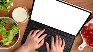 Sağlıklı bir kadın sağlıklı bir kahvaltı yaparken tablet bilgisayarında çalışıyor. Salata kasesi ve taze domatesler masada. en üstteki görünüm