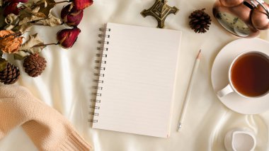 Üst manzara, arka planı boş spiral defter, çay fincanı, klasik saat, kurumuş çiçekler ve beyaz arka planda süveter olan kadınsı çalışma alanı. Kış, sonbahar kompozisyonları