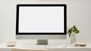 Bilgisayarlı, beyaz ekranlı, klavyeli, fareli, kahve fincanlı, not defterli, beyaz duvarda çiçek vazosu olan minimal ofis çalışma alanı. yakın plan