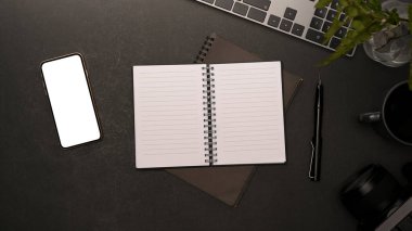 Boş ekran akıllı telefonlu en küçük iş yerinin üst görünümü, defter kağıdına kopyalama alanı ve siyah arkaplan üzerine ofis malzemeleri.