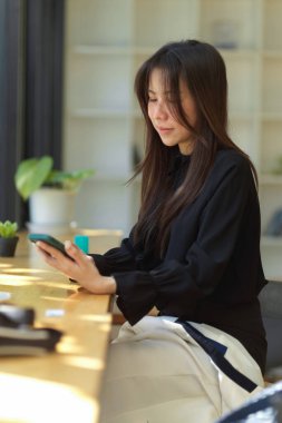 Kafedeki sehpada oturan ve akıllı telefonuyla yazı yazan siyah elbiseli Asyalı genç bir kadının portresi..