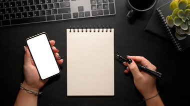 Cep telefonu ekranında bilgi arayan ve modern karanlık ofis masasındaki sarmal not defterine not alan bir kadın. Yakın çekim, üst görünüm