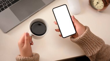 Düz yatıyordu, Top view 'de, sabah kahve fincanı ve akıllı telefonunu çalışma alanının üzerinde tutan örgü örülü süveter giymiş bir kadın. akıllı telefon boş ekran modeli.