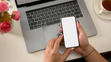 Üst çekim, beyaz arka planda dizüstü bilgisayarı boş ekranlı akıllı telefon kullanan kadın elleri. Düz yatıyordu