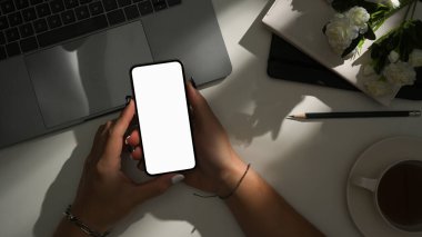 Üst çekim, modern hipster çalışma alanı. Kadın eli akıllı telefon boş ekran maketi ve güneş ışığı gölgesi tutuyor..