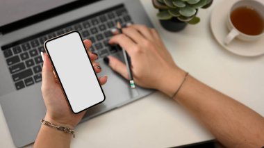 Dizüstü bilgisayarda çalışan, klavyede yazan ve elinde akıllı telefon modeli tutan bir kadın memur. en üstteki görünüm