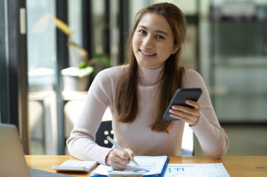 Akıllı telefon tutan ve ofis iş yerindeki mali raporlar üzerinde çalışan çekici bir kadın işçi ya da finans asistanı.