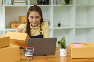 Güzel, Asyalı genç bir bayan iş sahibi dizüstü bilgisayardaki müşteri adresine bakıyor ve küçük bir paket kutusuna yazı yazıyor. E-ticaret, Nakliye, Girişim işi, Çevrimiçi alışveriş işi.