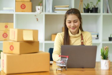 Teslimat paketleriyle dolu bir odada dizüstü bilgisayar üzerinde çalışan Asyalı çekici bir bayan girişimci ya da çevrimiçi bir iş sahibi. KOBİ iş ve e-ticaret kavramı