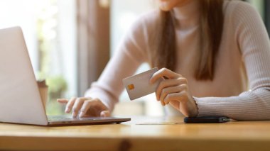 Dizüstü bilgisayar kullanan ve çalışma alanında kredi kartı tutan genç bir kadının kırpılmış, yakın plan görüntüsü. Çevrimiçi ödeme, internet bankacılığı, para transferi.
