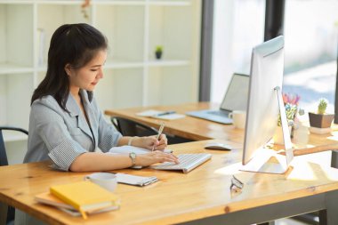 Resmi takım elbiseli güzel bir yönetici kadın yönetici, modern ofiste oturuyor, bilgisayar üzerinde çalışıyor ve not defterine not alıyor. Ofiste çalışan iş kadını.