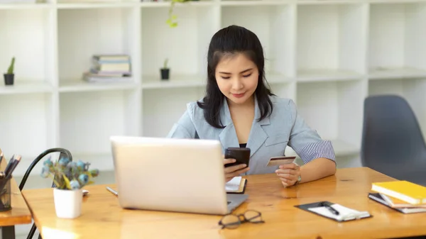 女性マネージャーは スマートフォン上のクレジットカードで彼女のオンラインショッピングを支払う 現代のオフィスに座っている — ストック写真