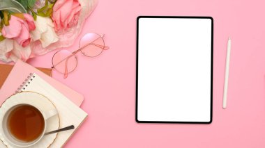 Ofis masasında dijital tablet beyaz ekran maketi, stilus kalem, gözlük, çay fincanı, not defteri ve pembe arka planda gül buketi. Üst görünüm, düz uzanma