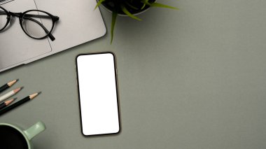 Afişinizi kırtasiye, dizüstü bilgisayar, kahve fincanı, bardaklar ve gri arkaplanda kopyalama alanı ile göstermek için bir akıllı telefon beyaz ekran modeli.