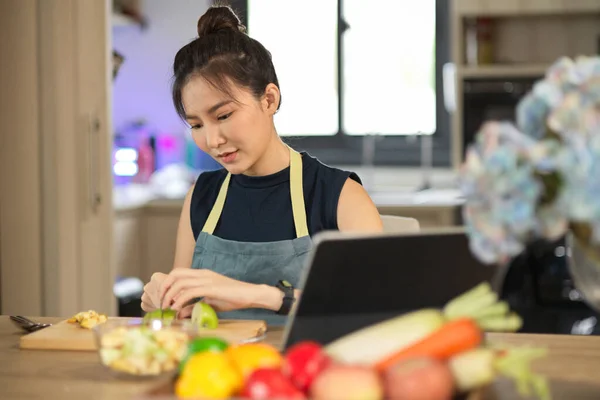 魅力的なアジアの若い女性は 台所のまな板にリンゴを切り刻みながら ポータブルタブレットを介してオンラインで健康的な食べ物を調理することを学ぶ — ストック写真