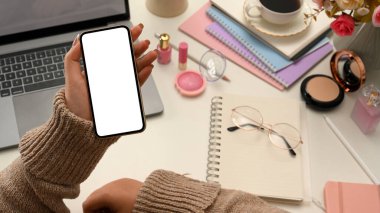 Milenyum modasına uygun, rahat kazaklı bir kadın çalışma masasında akıllı telefonuyla bazı online makaleler okuyor. telefon beyaz ekran modeli.