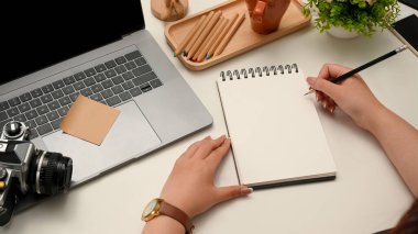 Kadın seyahat blogcusu, seyahat yorumlarını modern ofis masasındaki sarmal not defterine yazıyor. Laptop, kamera ve aksesuarlar masaya..