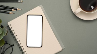 Akıllı telefon beyaz ekran maketi, spiral not defteri, kahve fincanı, kalemler, gri arka planda dekor olan modern minimal çalışma alanı. en üstteki görünüm