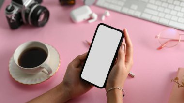 Yan görünüş, yakın plan, akıllı telefon beyaz ekran modelini şık pembe ofis masasının üzerinde tutan kadın elleri..