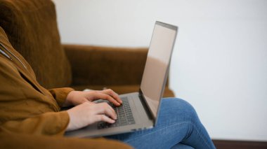 Yan görüş, kırpılmış çekim, rahat koltukta oturan ve evde dizüstü bilgisayarda çalışan bir kadın..