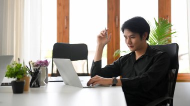 Siyah gömlekli Asyalı genç işadamı modern ofiste oturuyor ve dizüstü bilgisayarla çalışıyor..