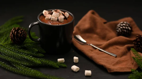 マシュマロ 松ぼっくり スプーン 茶色のテーブルクロスと黒の背景に緑の葉で溶かしたホットチョコレート — ストック写真