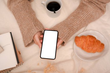 Tepeden çekilmiş, yemek masasının üzerinde akıllı bir telefon tutan kadın sosyal medya platformunda vakit geçiriyor. Telefon boş ekran modellemesi.