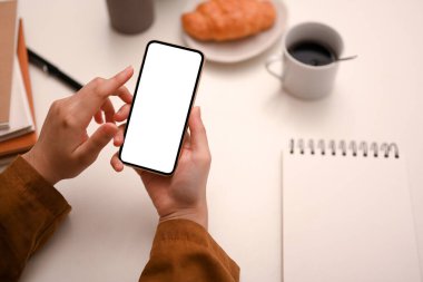 Tepeden çekim, ofis masasında kahvaltı ederken modern akıllı telefon kullanan kadın elleri. telefon beyaz ekran modeli.