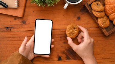 Üst manzara, kadın çalışma alanında rahatlıyor, kurabiye yiyor ve ahşap masasının üzerinde akıllı telefon kullanıyor..