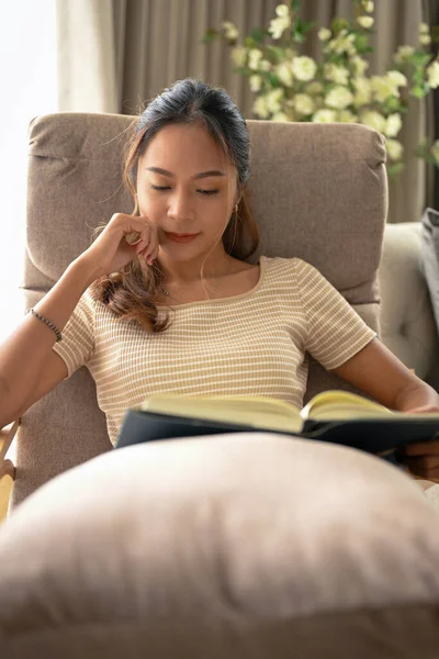 松懈的亚洲年轻漂亮女人坐在舒适的扶手椅上 在客厅里读一本小说 — 图库照片