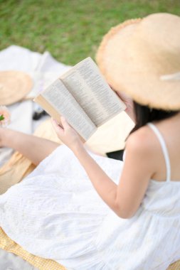 Yakından çekilmiş, beyaz elbiseli, en sevdiği romanı okuyan, yeşil arka bahçede pikniğin tadını çıkaran genç bir kadın..