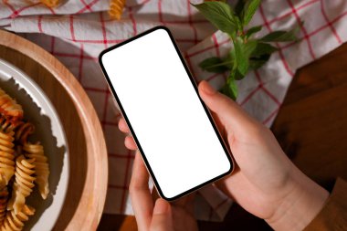 Yakın çekim, üst manzara, akıllı telefon tutan kadın elleri yemek masasının üzerinde makarna ile beyaz ekran modeli.