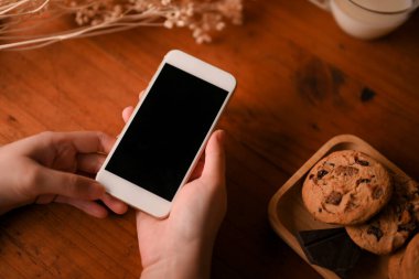 Bir tabak kurabiye ve dekor ile ahşap masanın üzerinde akıllı telefon taşıyan bir kadın..