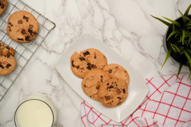 Beyaz bir tabakta çikolatalı kurabiyeler, bir bardak süt, taze pişmiş kurabiyeler ve mermer arka planda dekor. Pastane, pasta konsepti