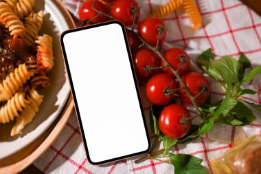 Akıllı telefon beyaz ekran modeli bir tabak makarna, taze domates ve fesleğen yaprağıyla birlikte bir yemek masasının üzerinde. İtalyan makarna mutfağı..