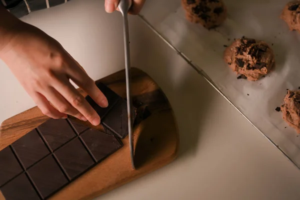 トップビュー 木製のまな板にチョコレートバーを切断ナイフを使用して女性の手 チョコレートクッキーを焼くための成分を準備します — ストック写真
