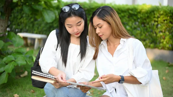 Çekici Asyalı kız üniversite öğrencisi parkta oturuyor, tablet ekrandan detaylara bakıyor ve arkadaşına tavsiyelerde bulunuyor..