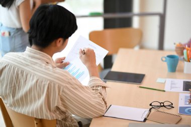 Arka tarafta, iş adamı toplantıda oturmuş, iş arkadaşının planı sunuşunu dinlerken elinde bir mali rapor tutuyor..