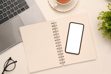Üst görünüm, akıllı telefon beyaz ekran modeli ile modern çalışma alanı boş spiral defter, dizüstü bilgisayar, gözlükler ve beyaz masa üzerindeki dekor.
